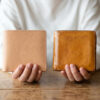 新しいライトタンのイルビゾンテ財布と、数年使い込まれて深い飴色にエイジングしたイルビゾンテ財布を並べて持つ日本人女性の手元。革製品の経年変化の魅力を伝える画像。