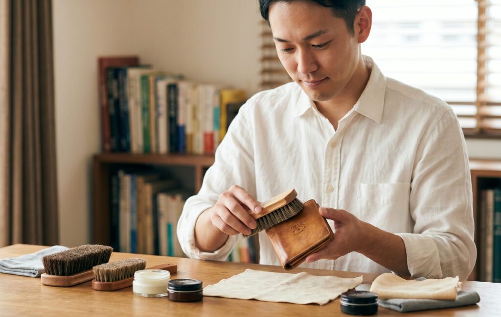 落ち着いた趣味の部屋で、日本人男性が穏やかな表情で、長年愛用して味わい深くなったイルビゾンテの財布に、馬毛ブラシとケアクリームを使って丁寧にメンテナンスを行っている様子。怖さをなくし、愛情を持ってケアする姿を伝える。