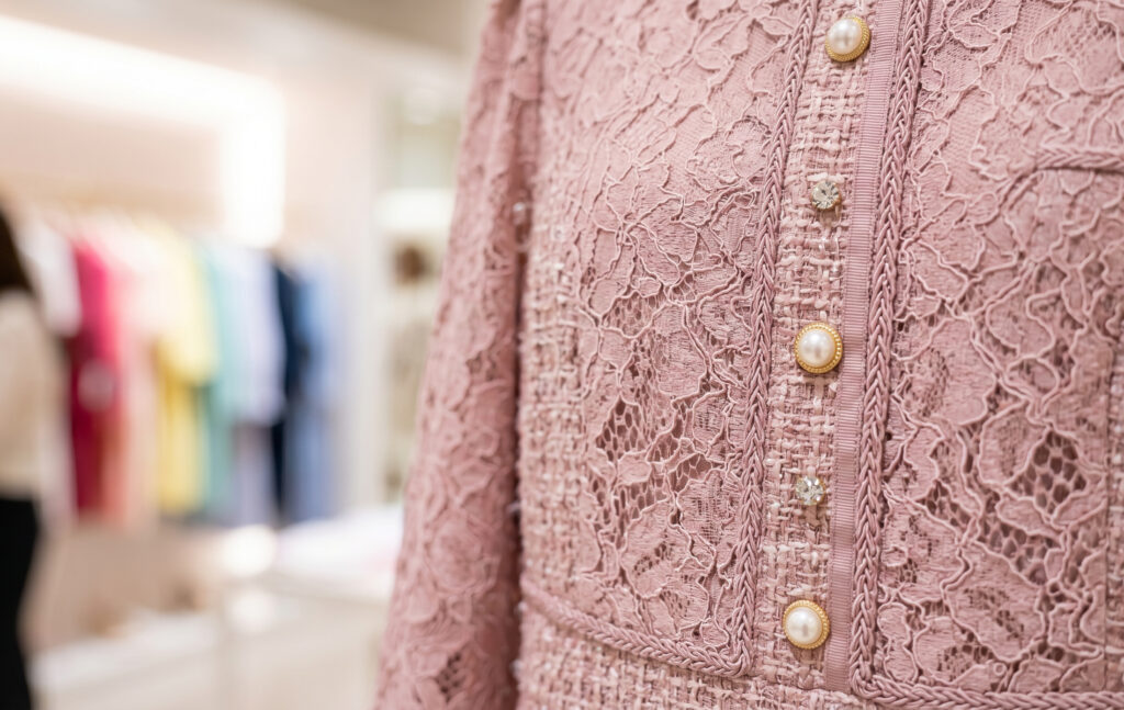 明るいブティックで、レースやツイード素材のワンピースに施されたパールボタンやビジューなどの装飾的なディテールのアップ写真。ブランドのこだわりを表現