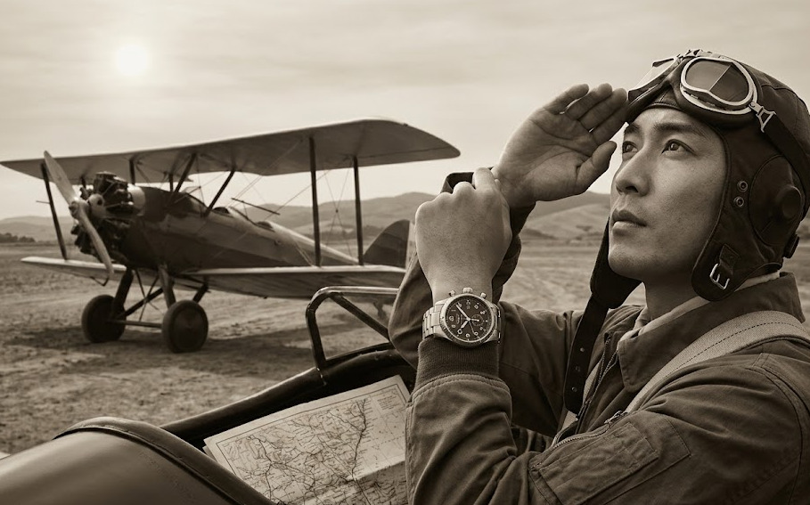 1920年代の飛行場を背景に、歴史的なアワーアングルウォッチを腕に着用した日本人パイロットが複葉機の横で空を見上げる。セピア調のヴィンテージ写真
