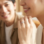 日本人の若いカップルが笑顔で婚約指輪を見つめる、幸せあふれる瞬間を捉えた写真。プラチナとダイヤモンドのリングが光る。