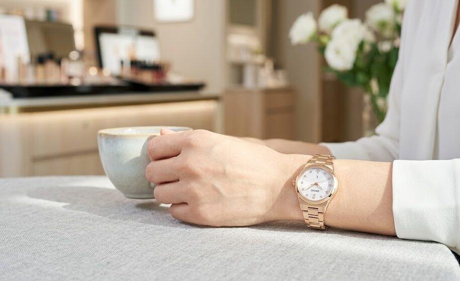 落ち着いたブティックラウンジで、50代の日本人女性が、自身の腕元のセイコールキア（ピンクゴールド）と、上品な真珠のネックレスを合わせている。時計は上品な輝きを放ち、手頃な価格ながら大人の女性を満足させる高級感があることを示唆している。