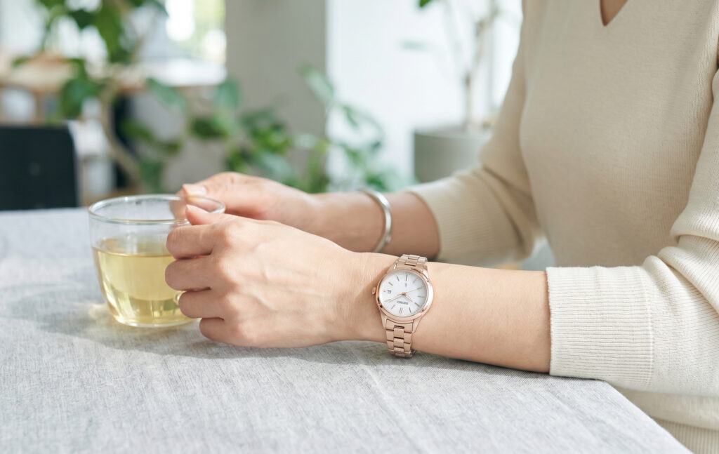 現代的なカフェで、日本人女性が腕元のセイコールキアの時計を確認している。彼女はベージュのニットを着用しており、時計はピンクブラウンの文字盤とステンレスのブレスレットで、大人の女性の日常に馴染む洗練されたデザイン。