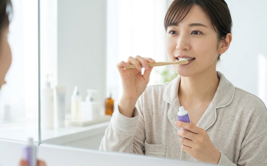 洗面台で、日本人女性がリラックスして、自然な動作で、片手に歯ブラシ、もう片手に無地のチューブを持ち、歯ブラシにジェルを付けている。副作用の心配がない安心感のあるケア風景。ロゴなし