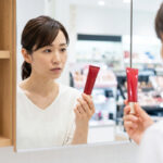 アスタリフトの赤いBBクリームを手に、不思議そうな表情で鏡を見る日本人の女性