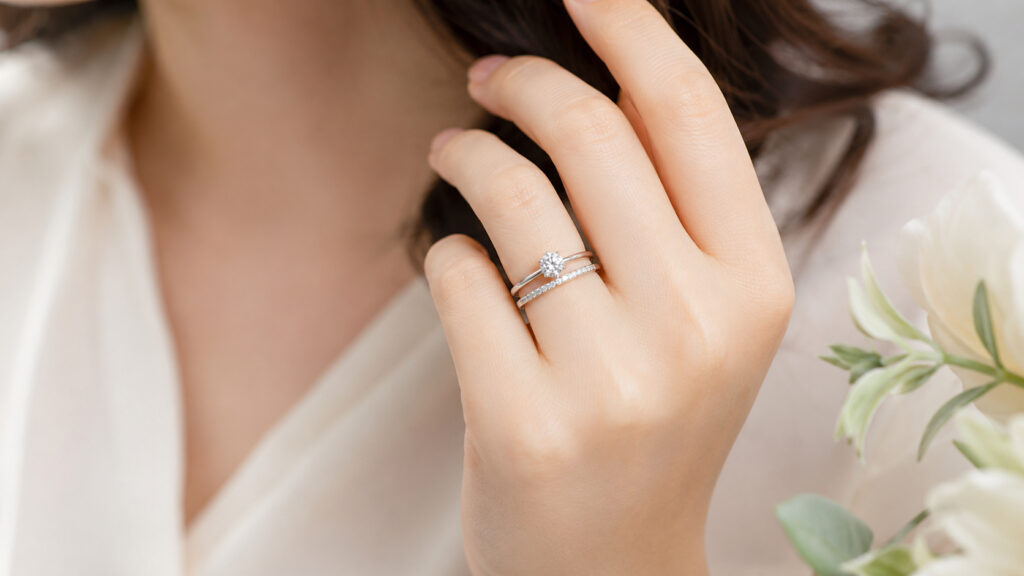 自然光の中、婚約指輪と結婚指輪を美しく重ね付けし、満足感と幸せに満ちた表情で指輪を見つめる日本人女性の手元。アーカー独自の「重ね付け」を視覚化。