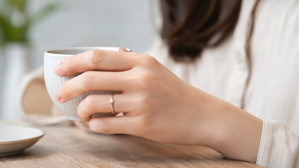 カフェで婚約指輪をつけてくつろぐ日本人女性。彼女の左手にはアーカーの華奢な婚約指輪が自然に馴染んでいる。日常使いでの強度と美しさを暗示