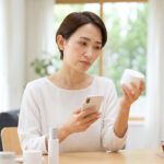 シミ対策のオールインワンジェルを手に取り、スマートフォンの画面を見ながら不安げに考える日本人女性