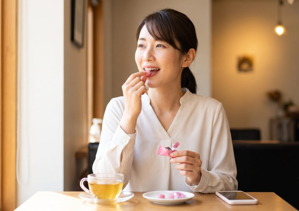 カフェでリラックスしている日本人女性が、ドモホルンリンクルのど飴を1粒口に入れ、満足げに微笑んでいる。手元には個包装の飴がある