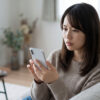 自宅のソファで、不安げな表情でスマホの画面を見つめる日本人女性