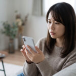 自宅のソファで、不安げな表情でスマホの画面を見つめる日本人女性