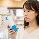 ドラッグストアの化粧品売り場で、ラロッシュポゼの日焼け止めを手に取り、価格を見て少し驚いた表情を浮かべる日本人女性