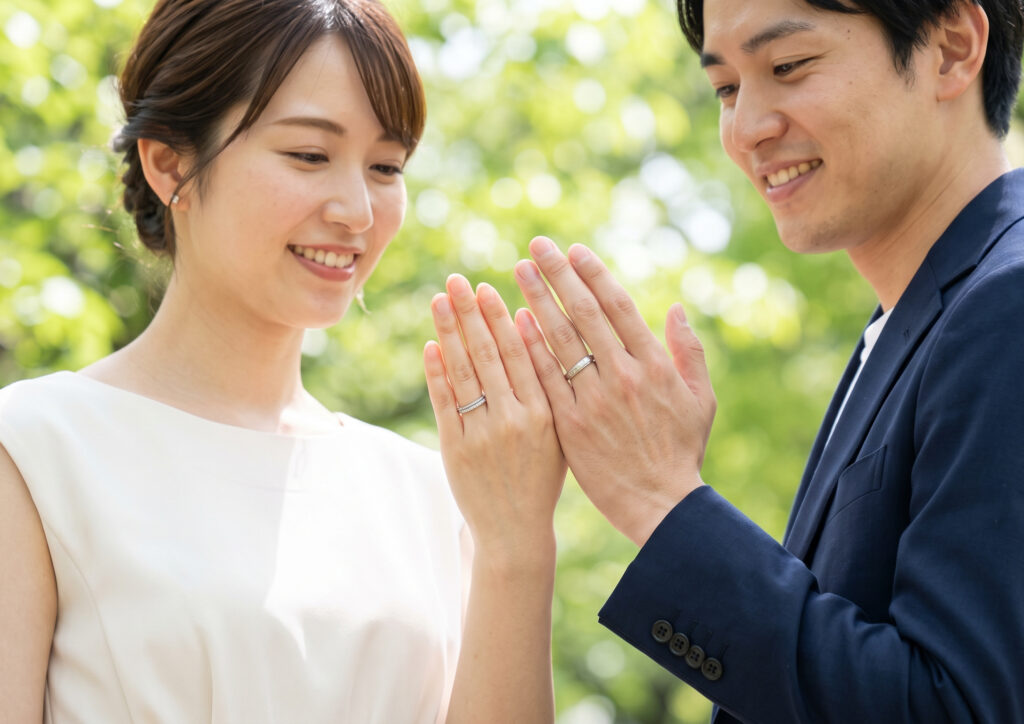 ダイヤ入りの結婚指輪をつけた日本人女性と、シンプルな結婚指輪をつけた日本人男性が、幸せそうに手を繋いでいる。屋外のパークにて、自然光のもとで