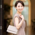 結婚式場のエントランスで、上品なパーティードレス姿で高級感のあるバッグ（文字なし）を持って笑顔で立つ日本人女性